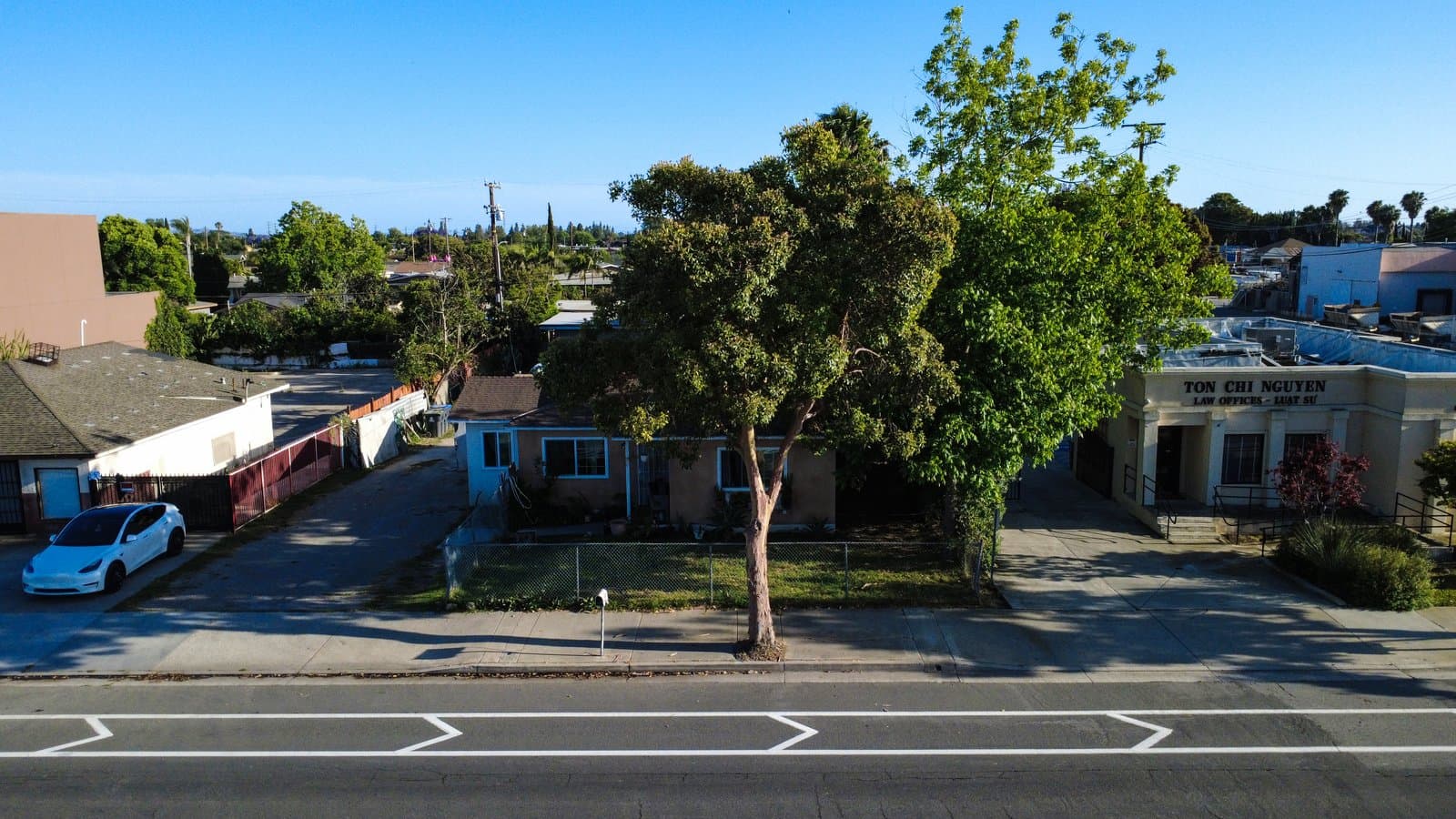 Aerial view of a commercial street featuring a law office building, a residential house, a white car, and numerous trees under a clear sky.