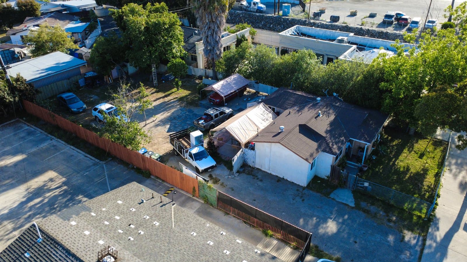 Aerial view of a residential neighborhood featuring several houses, lush green trees, various parked cars, and fences, adjacent to a large commercial building with a flat roof and a paved parking lot.