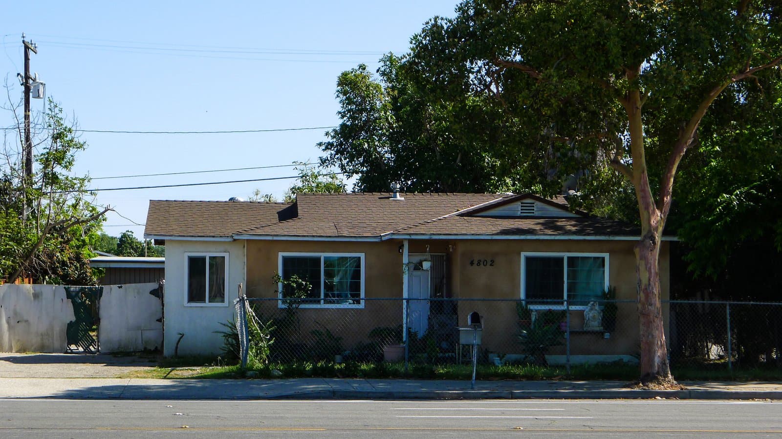 A tan single-story house with a brown roof and white trim, featuring the address 4802, is visible behind a chain-link fence. The house is viewed from a paved street with double yellow lines and a sidewalk. A large tree stands to the right of the house, and a utility pole is visible on the left against a clear blue sky.